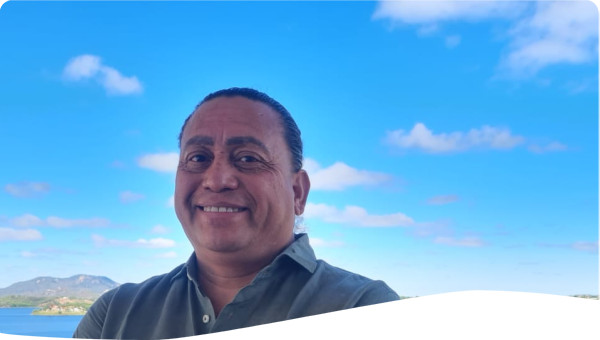 Em primeiro plano, um homem sorridente de braços cruzados veste camisa verde e está em uma varanda. Ao fundo, uma paisagem do Semiárido brasileiro com um lago azul, vegetação verde-amarelada e montanhas no horizonte sob céu azul com nuvens esparsas