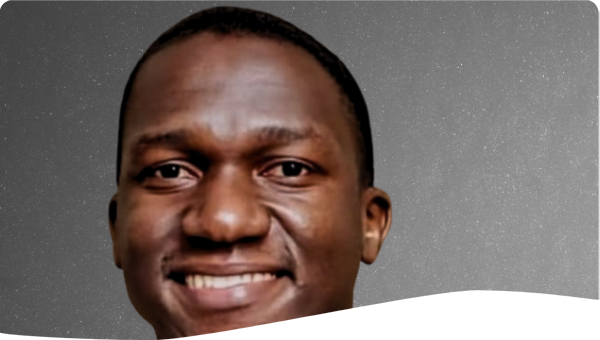 A man wearing a dark blue suit, patterned tie, and pocket square, smiling against a plain dark background.