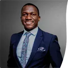 A man wearing a dark blue suit, patterned tie, and pocket square, smiling against a plain dark background.
